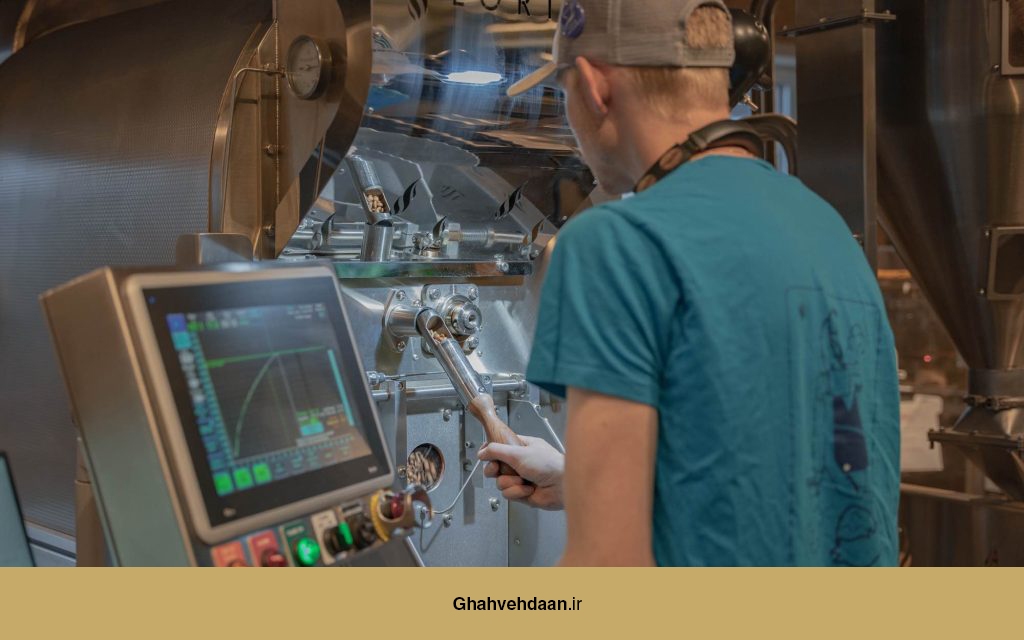 یک رُستر قهوه در حال بررسی دانههای رُستشده در کنار دستگاه رُست پیشرفته با صفحه کنترل دیجیتال.