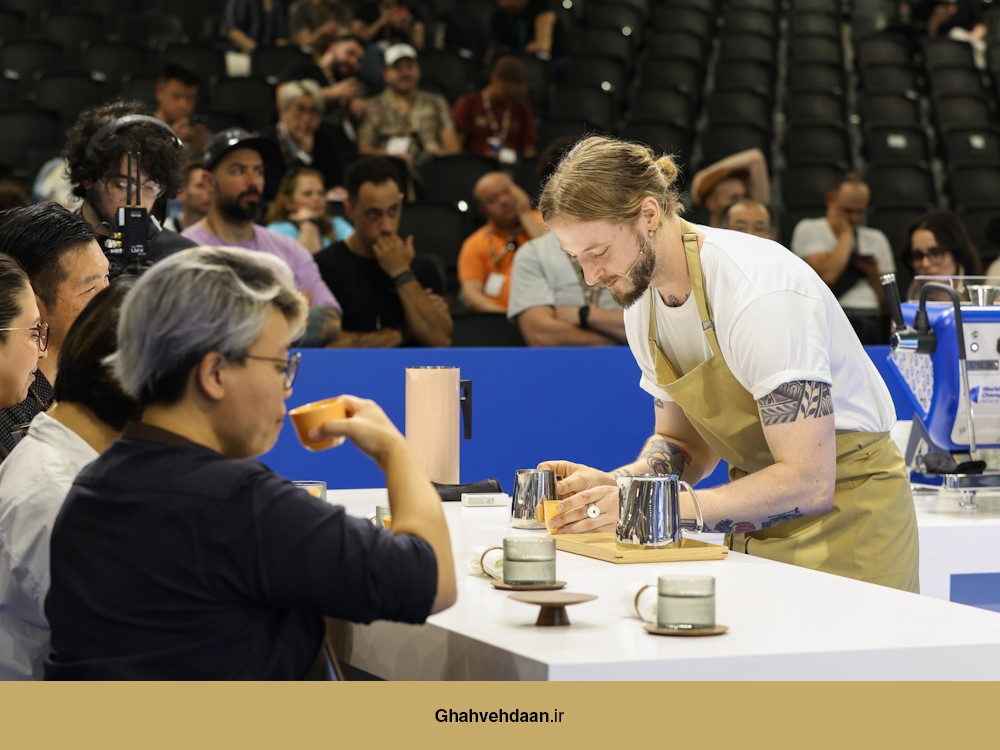 پاتریک رولف، باریستای شرکتکننده در مسابقات قهرمانی جهانی باریستا، در حال آمادهسازی نوشیدنی برای داوران، در یک محیط رقابتی با تماشاگران در پسزمینه.